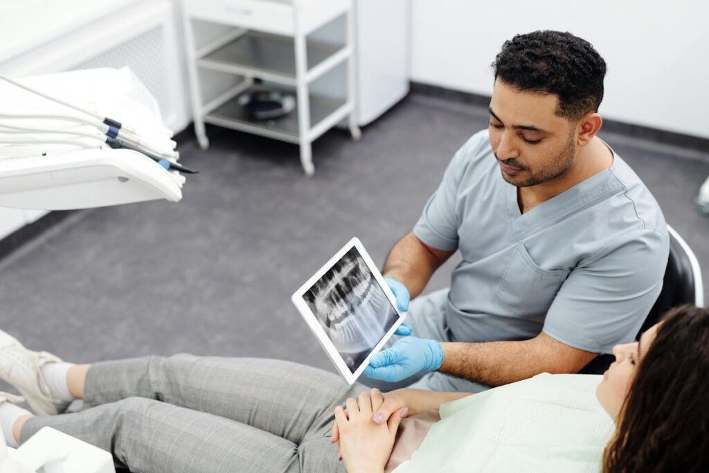 Man in Gray Scrub Suit Sitting Beside a Patient while Holding Digital Tablet