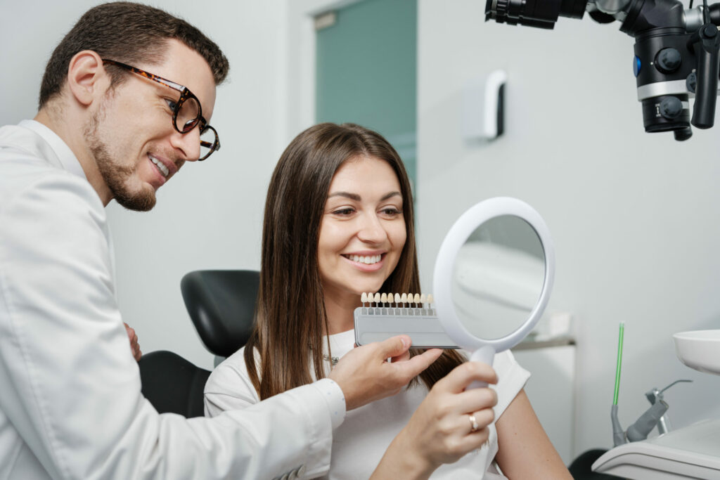 Young male dentist in white coat show veneers enamel color palette for beautiful patient woman sitting in a dental unit. Healthcare oral aesthetic treatment.