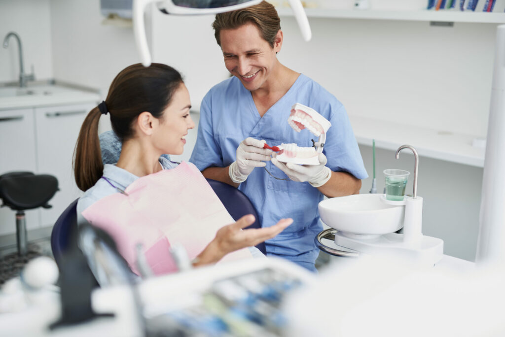 Consultation. Portrait of beautiful lady sitting in dental chair while stomatologist in sterile gloves holding false teeth and toothbrush