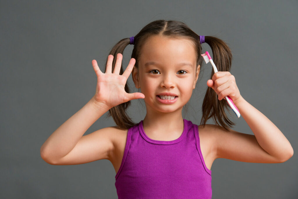 Child showing number five with her hand and fingers while holding a toothbrush on the other hand.